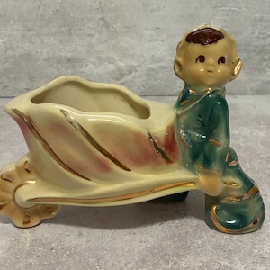 May include: A vintage ceramic planter in the shape of a wheelbarrow, featuring a seated child figure. The planter is cream-colored with pink and gold accents. The child figure is dressed in green with gold trim.