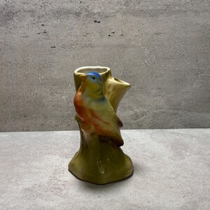 May include: A ceramic vase shaped like a tree stump with a bird perched on the side. The bird features a blue head, yellow neck, and orange chest. The tree stump base is colored in shades of olive green and brown with a textured, bark-like surface.
