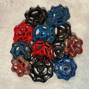 May include: A collection of twelve metal valve handles in various colors including red, blue, black, and brown. Each handle features a circular, multi-spoked design with a square center opening. The surfaces show signs of wear, paint chipping, and rust.
