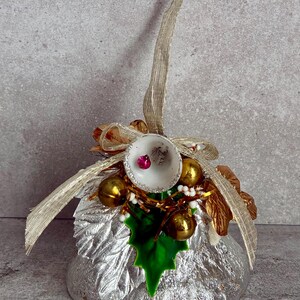 May include: A silver-colored Christmas ornament with gold accents. The ornament features a bell-shaped design with a silver base, adorned with gold bells, a green holly leaf, and a gold ribbon. A pink gem is inside the bell.