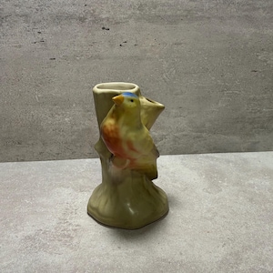 May include: A ceramic vase shaped like a tree stump with a yellow and orange bird perched on the side. The bird has a small blue patch on its head and a yellow beak. The vase features a textured, light green finish and a hollow opening at the top.