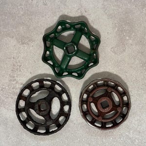 May include: Three metal valve handles arranged in a triangular formation. The top handle is painted green with a scalloped edge. The bottom left handle is dark, textured, and rusted. The bottom right handle is a smooth, solid brown color.