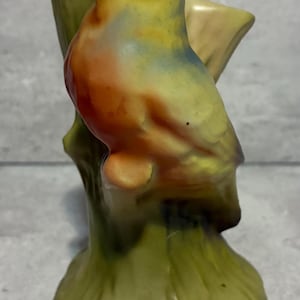 May include: A ceramic bird figurine perched on a tree branch. The bird has a yellow breast, orange belly, and blue head. The base is green, and the branch is a light tan color. The figurine is a decorative item.