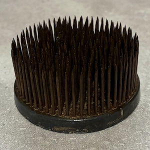 May include: A round, dark brown flower frog with numerous sharp metal pins. The pins are densely packed and protrude from a circular base. The base is a dark color, possibly black, and shows signs of wear.