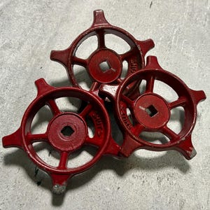 May include: Three circular red metal valve handles with five spokes each. The center of each handle features a square opening for attachment. The word SHUT is embossed on one of the spokes of each handle, with a directional arrow pointing clockwise.