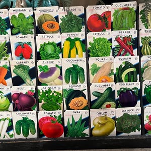 May include: A wooden display shelf with 36 vintage seed packets. The packets are arranged in four rows of nine. Each packet features a colorful illustration of the vegetable or flower it contains. The packets are labeled with the name of the plant and the price. Some of the plants pictured include: tomato, lettuce, cucumber, pepper, squash, okra, radish, and onion.