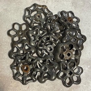 May include: A collection of antique, metal water valve handles. The handles are various shapes, including flower and gear designs. The metal has a dark, aged appearance with some rust visible, suggesting age and use. The handles are arranged in a pile.