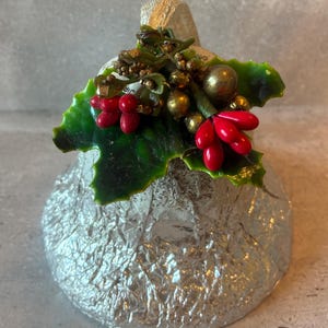 May include: A silver-colored bell ornament with a textured surface. The bell is adorned with green leaves, red berries, and gold-colored accents, creating a festive holiday decoration.