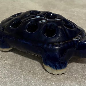 May include: A ceramic turtle-shaped flower frog in a deep blue glaze. The turtle has multiple holes on its back for arranging flowers. The feet are a contrasting off-white color. A decorative item for floral arrangements.
