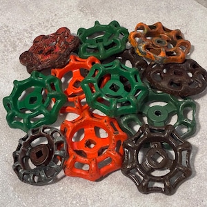 May include: A collection of twelve metal valve handles in various colors including green, orange, brown, and rusted red. Each handle features a circular, multi-spoked design with a square center opening, showing signs of wear and weathered paint.
