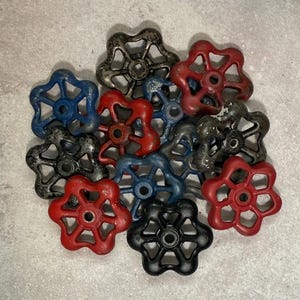 May include: A collection of metal valve handles in various colors including red, blue, black, and weathered gray. Each handle features a five-spoke star design with a central circular opening, showing signs of wear and paint chipping on the surfaces.