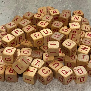 May include: A collection of light brown wooden cubes featuring red letters and small numerical values printed on each face. These square game pieces are scattered in a pile, displaying various characters such as Q, U, D, T, R, and F.