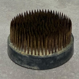 May include: A round, dark gray flower frog with numerous bronze-colored pins. The pins are closely spaced and arranged in a circular pattern, rising from the base. The base is a dark gray color, and the overall design is simple and functional.