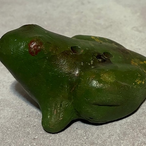 May include: A green frog figurine with a slightly distressed finish. The frog has a small red spot on its head and yellow accents. The figurine is made of a solid material and has a smooth, rounded shape. A decorative item.