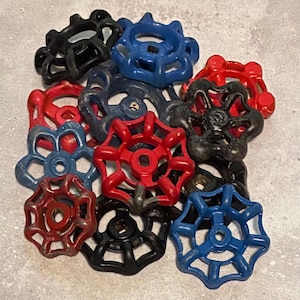 May include: A collection of vintage water valve handles in various colors, including red, black, and blue. The handles have a starburst design and are made of metal. The image shows a close-up of the handles, with some wear and tear visible.