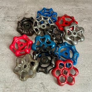 May include: A collection of vintage valve handles in various colors, including red, blue, black, and silver. The handles have a star-shaped design with a square center hole. These industrial-style handles are likely made of metal and show signs of wear.