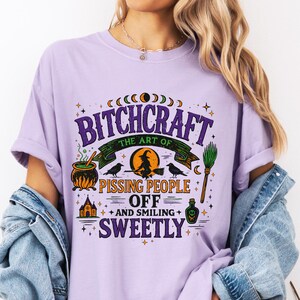 May include: Orchid-colored t-shirt with the text "BITCHCRAFT the art of pissing people off and smiling sweetly" in a whimsical design. The graphic includes a cauldron, witch, crows, and a broom.