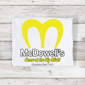 May include: White t-shirt featuring a yellow "M" logo, with the text "McDowell's Home of the Big Mick!" in black and yellow. The text "Queens, New York" is printed below the logo. The shirt is folded.