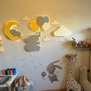 Könnte beinhalten: Ein Kinderzimmer mit einer weißen Wand, die mit einem Mond, Sternen, Wolken und einem Elefanten, der Ballons hält, dekoriert ist. Auf einem Regal stehen Stofftiere, darunter eine Giraffe, eine Raupe und eine Robbe. Es gibt einen kleinen Tisch mit einem Buch und Buntstiften darauf.