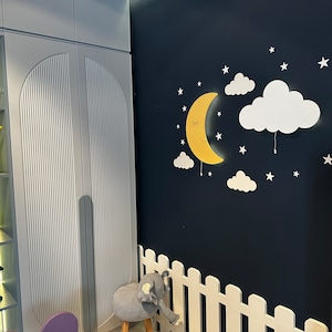 May include: A white wooden bookshelf with a white picket fence and a yellow crescent moon wall light with clouds. The wall is painted a dark blue color.