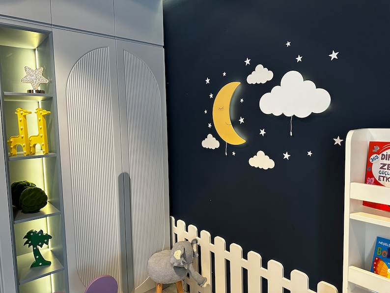 May include: A white wooden bookshelf with a white picket fence and a yellow crescent moon wall light with clouds. The wall is painted a dark blue color.