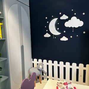 May include: A white picket fence surrounds a small table and chairs in a playroom. The wall is painted a dark blue and features a white moon and cloud nightlight with stars. A bookshelf with colorful books is visible in the background.