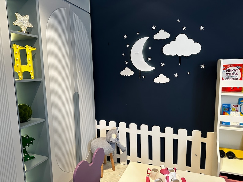 May include: A white picket fence surrounds a small table and chairs in a playroom. The wall is painted a dark blue and features a white moon and cloud nightlight with stars. A bookshelf with colorful books is visible in the background.
