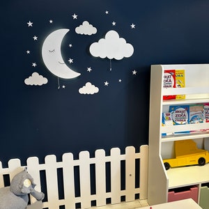 May include: White wooden fence with a white moon and cloud wall decor, a stuffed elephant toy, and a white bookshelf with colorful books and a yellow toy truck.
