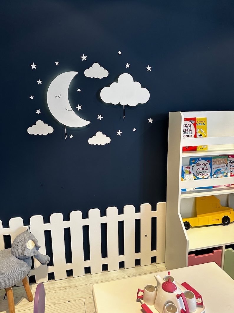 May include: White wooden fence with a white moon and cloud wall decor, a stuffed elephant toy, and a white bookshelf with colorful books and a yellow toy truck.