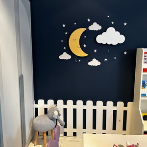 May include: A white picket fence with a gray elephant stool in front of it. A yellow crescent moon with a smiling face and closed eyes is on a dark blue wall with white clouds and stars. A white bookshelf with books in Turkish is on the right side of the image.