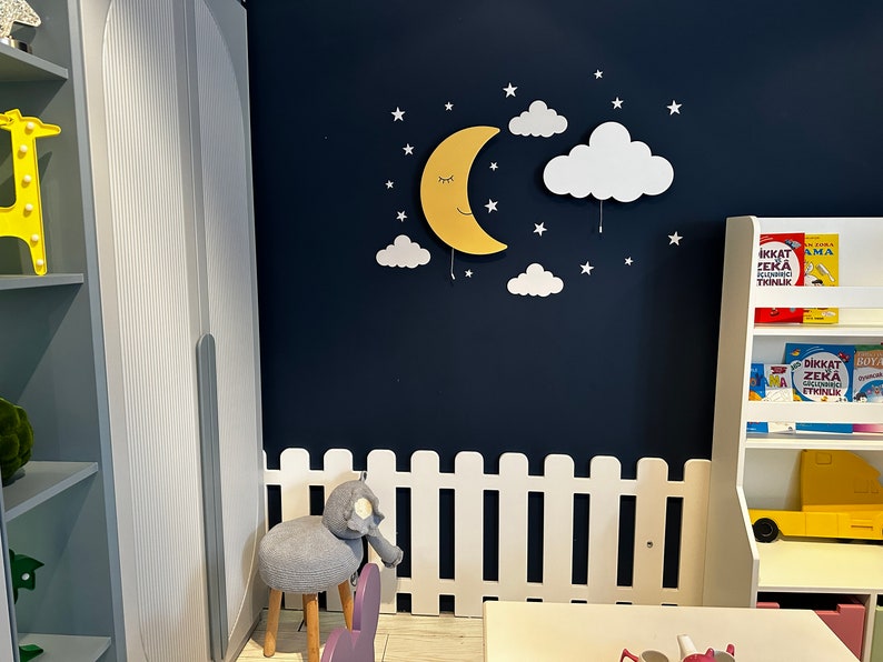 May include: A white picket fence with a gray elephant stool in front of it. A yellow crescent moon with a smiling face and closed eyes is on a dark blue wall with white clouds and stars. A white bookshelf with books in Turkish is on the right side of the image.