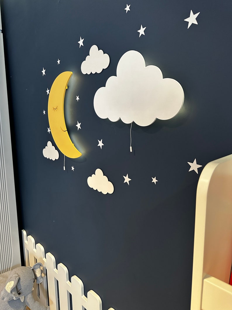 May include: A yellow crescent moon and white cloud wall decor with a string of white stars on a dark blue background. The moon and cloud are 3D and have a light inside.