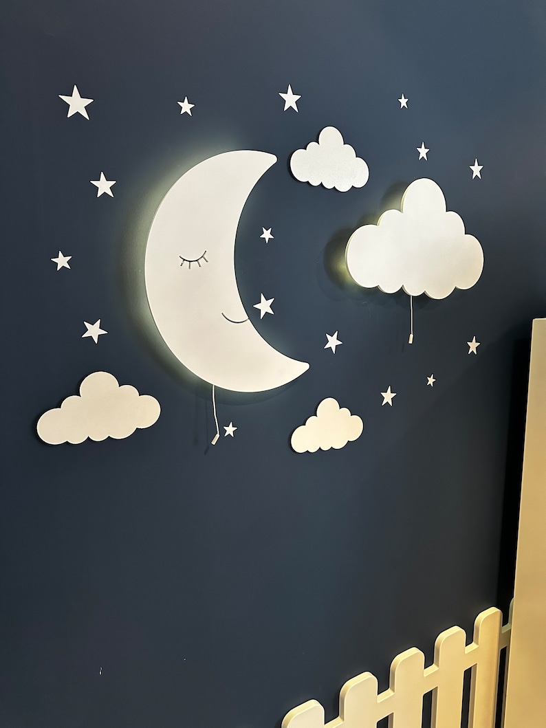 May include: A white crescent moon wall light with a smiling face and closed eyes, surrounded by white clouds and stars on a dark blue wall.
