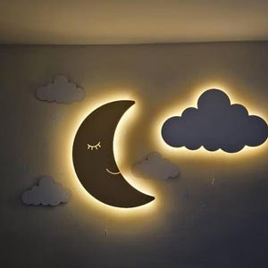 May include: A wall-mounted night light set featuring a crescent moon with a smiling face and a cloud, both outlined with a warm, yellow glow. Additional white cloud cutouts are placed around the moon and cloud. The set is designed for a nursery or children's room.