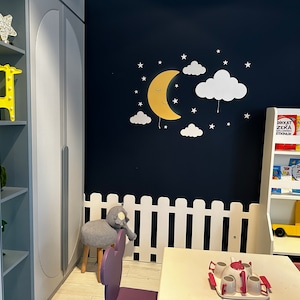 May include: A white picket fence surrounds a play area with a white table and chairs. A yellow toy truck is on a shelf with books that say "Dikkat Zeka Etkinlik" in Turkish. A yellow crescent moon with a smiling face and white clouds are on the wall.