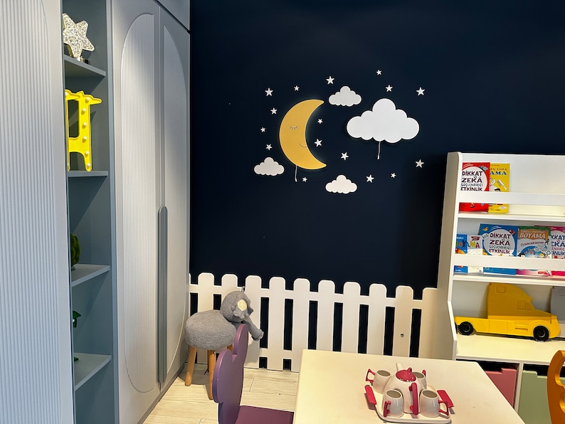 May include: A white picket fence surrounds a play area with a white table and chairs. A yellow toy truck is on a shelf with books that say "Dikkat Zeka Etkinlik" in Turkish. A yellow crescent moon with a smiling face and white clouds are on the wall.