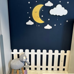 May include: A white picket fence with a gray knitted elephant stool in front of it. The wall behind the fence is painted a dark blue and features a yellow crescent moon with a smiling face and white clouds with stars.