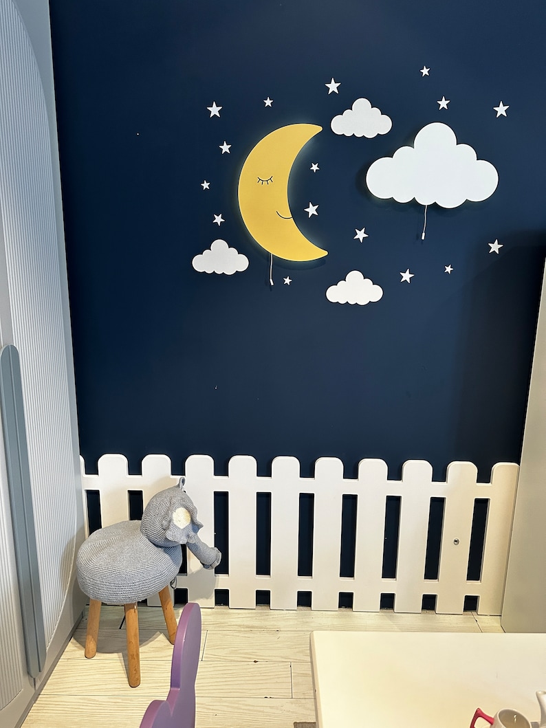 May include: A white picket fence with a gray knitted elephant stool in front of it. The wall behind the fence is painted a dark blue and features a yellow crescent moon with a smiling face and white clouds with stars.