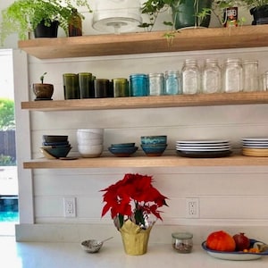 Puede incluir: Una cocina con estantes de madera que exhiben varios artículos. Los estantes contienen plantas, cristalería, cuencos y platos. Una flor de Pascua en una maceta dorada se encuentra en la encimera, junto con un cuenco, un tarro y un plato de fruta.