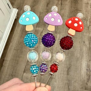 May include: Three decorative mushroom-topped skewers. The mushroom caps are light blue, purple, and red with white polka dots. Each skewer has a beaded design in coordinating colors, with a silver-colored metal stem. The mushroom tops are adorned with rhinestones.