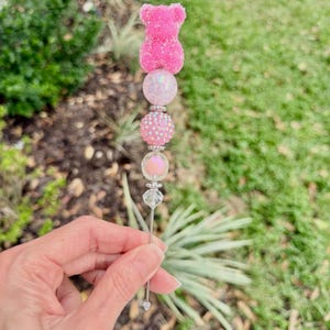 May include: A pink decorative skewer with a gummy bear topper, iridescent beads, and clear crystal accents. The skewer is held in a hand against a blurred green background.