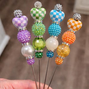 May include: A collection of decorative beaded pins with heart-shaped tops in various colors, including purple, green, blue, and orange. Each pin features a sparkling rhinestone accent and a silver-colored metal pin. The pins are held in a hand.