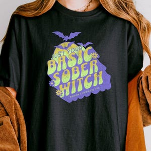 May include: Black t-shirt with the text "JUST YOUR BASIC SOBER WITCH" in neon green and purple. The graphic includes a bat and floral accents. The shirt is part of a clothing collection.