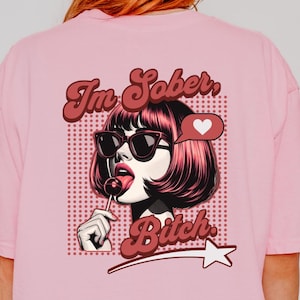 May include: A light pink t-shirt with a crew neck and short sleeves. The back of the shirt features a graphic of a woman with sunglasses and a lollipop, with the text "I'm Sober, Bitch."
