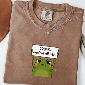 May include: Brown Comfort Colors t-shirt with the text "SOBER. against all odds." and a cartoon frog holding a sign. A silver necklace with a small pendant is draped across the shirt.