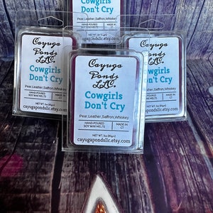 May include: Four purple wax melt containers with the text "Cayuga Ponds LLC. Cowgirls Don't Cry" and the scent description "Pear, Leather, Saffron, Whiskey". Each container is labelled "Hand-Poured Soy Wax Melts" and "Made in CT".