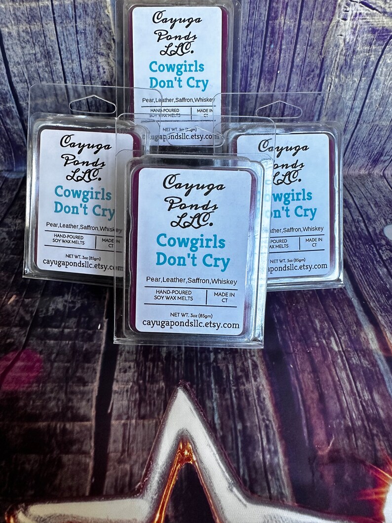 May include: Four purple wax melt containers with the text "Cayuga Ponds LLC. Cowgirls Don't Cry" and the scent description "Pear, Leather, Saffron, Whiskey". Each container is labelled "Hand-Poured Soy Wax Melts" and "Made in CT".