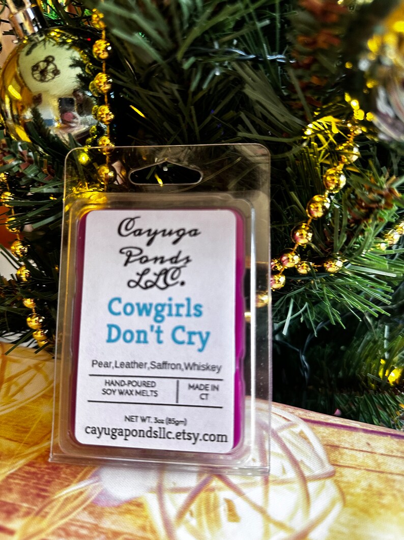 May include: A purple soy wax melt in a clear plastic container. The label reads "Cayuga Ponds LLC. Cowgirls Don't Cry Pear, Leather, Saffron, Whiskey Hand-Poured Soy Wax Melts Made in CT Net Wt. 3oz (85gm) cayugapondsllc.etsy.com"