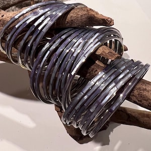 May include: A collection of silver bangles with a mother-of-pearl inlay. The bracelets are stacked on a piece of driftwood, creating a contrast between the cool metal and the warm wood tones. The bangles have a reflective surface.