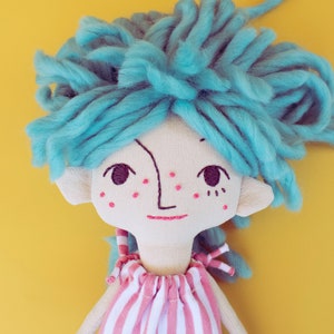 May include: A handmade doll with a whimsical design. The doll features a light-colored fabric face with embroidered details, including a simple smile and freckles. It has bright blue yarn hair and a striped pink and white dress. The background is a solid yellow.